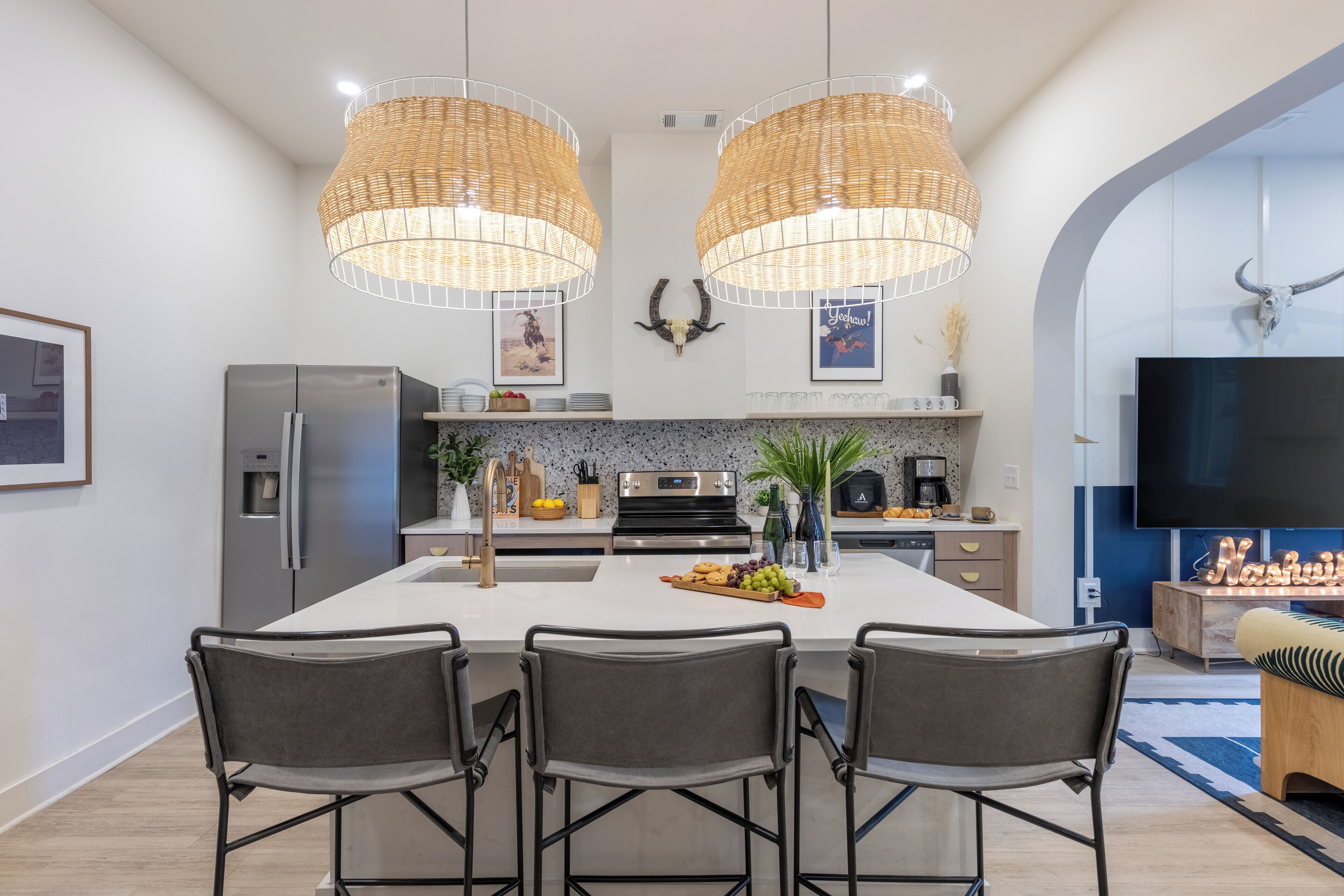 A modern kitchen with a central island, two pendant lights, a refrigerator, a stove, and a TV in the adjacent living area.