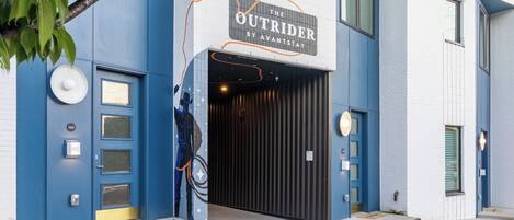 A building entrance with a sign reading 'The Outrider by Avantstay,' featuring a modern design with blue and white walls, a dark entrance door, and a tree visible in the foreground.