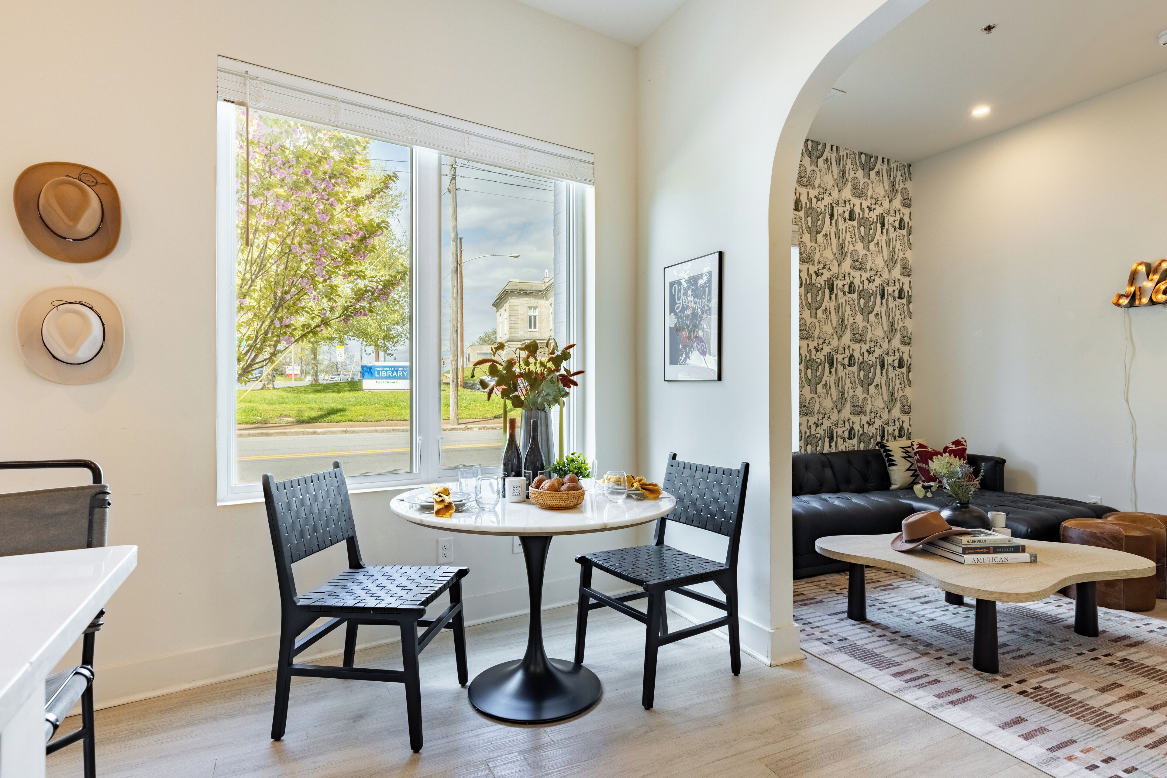 A modern living room with a round dining table, two chairs, a sofa, and a coffee table. A window offers a view of a tree and a building.