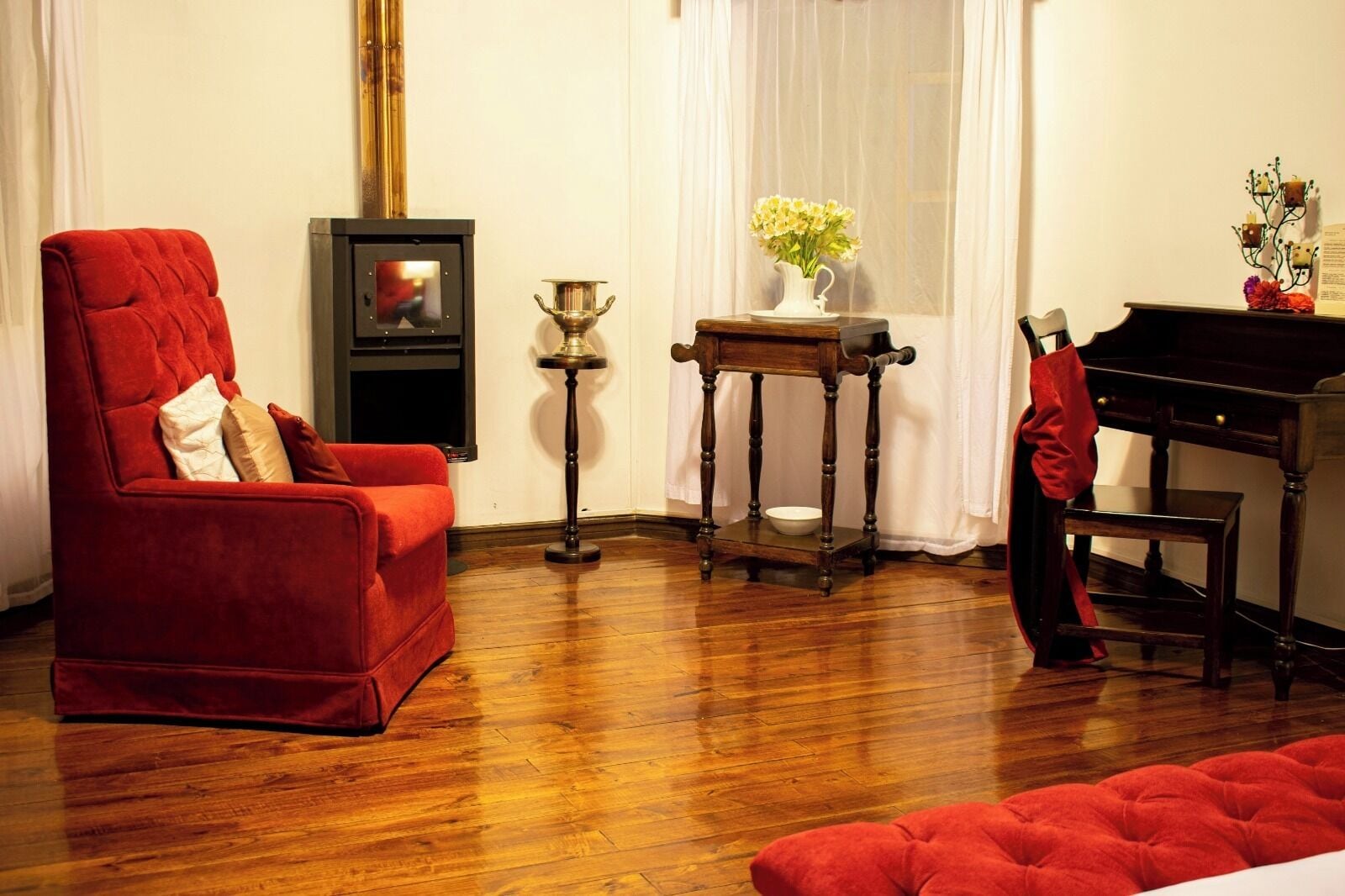 Una habitación con un sillón rojo, un piano de madera, una pequeña mesa con un jarrón de flores y una lámpara decorativa.
