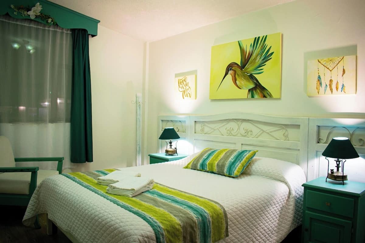 Een net opgemaakt bed met een gestreept kussen, een nachtkastje met een lamp, en een muur met een schilderij van een kolibrie.