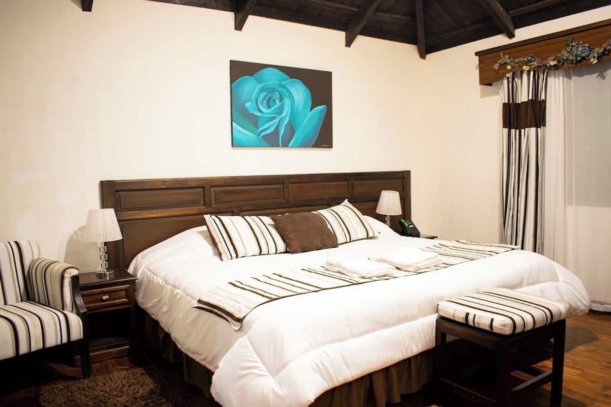 Een netjes ingerichte slaapkamer met een groot bed, een nachtkastje met een lamp, een stoel en een wanddecoratie van een blauwe roos.