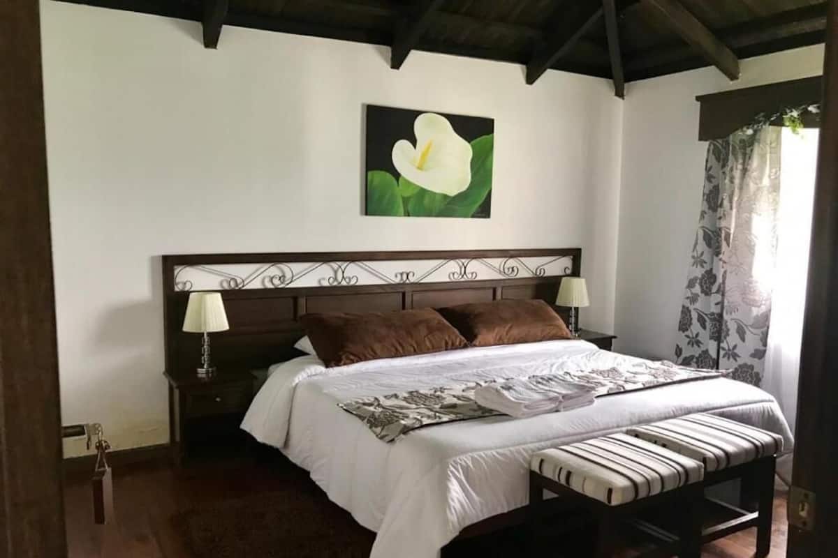 Een slaapkamer met een houten bed, wit beddengoed, een nachtkastje met een lamp en een bloemschilderij aan de muur.