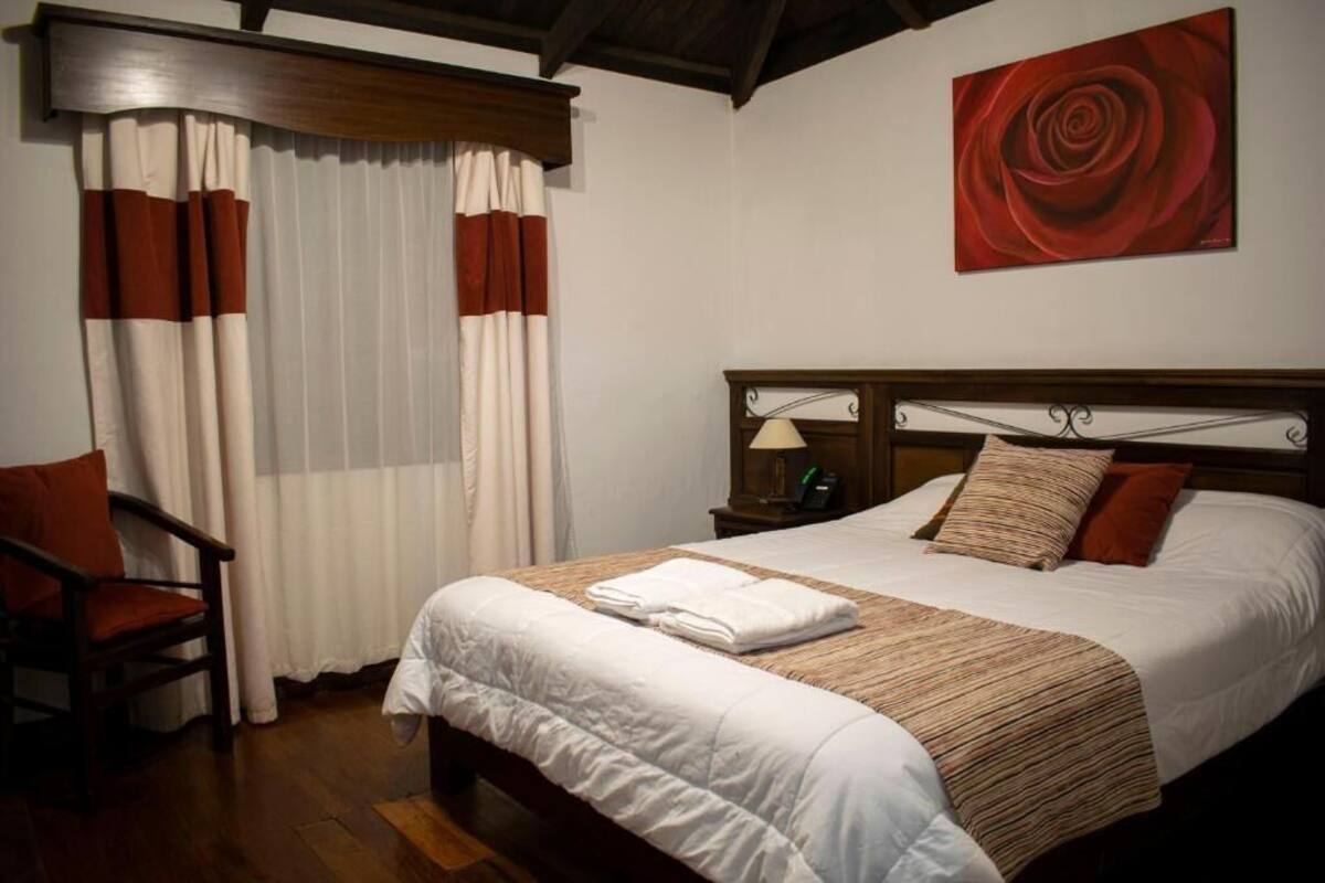 Een netjes opgemaakt bed met een wit-bruin gestreept dekbedovertrek, een houten nachtkastje met een lamp en een schilderij van een rode roos aan de muur.