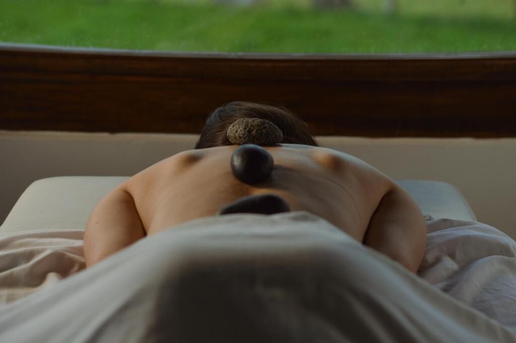 A person lying on a bed with a stone on their back, viewed from behind.