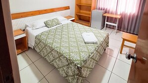 Habitación de hotel con cama, escritorio, silla, estante, ventilador y ventana con cortinas.