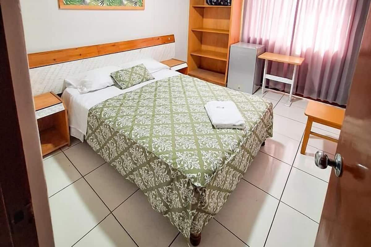 Hotelkamer met een bed, bureau, stoel, plank, ventilator en raam met gordijnen.
