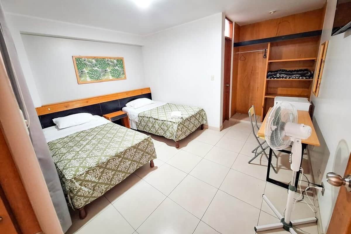 Een hotelkamer met twee bedden, een houten garderobe, een ventilator en een schilderij aan de muur.
