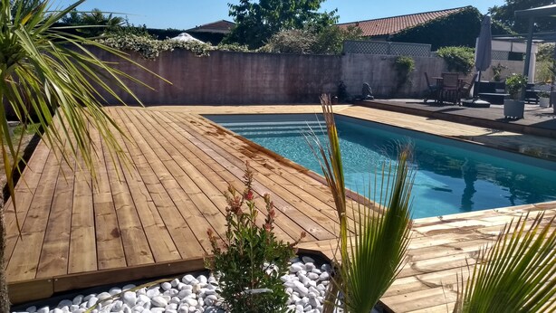 Eine Holzterrasse umgibt einen rechteckigen Pool mit einer Steinborduraus, im Vordergrund eine kleine Pflanze und im Hintergrund Gartenmöbel.