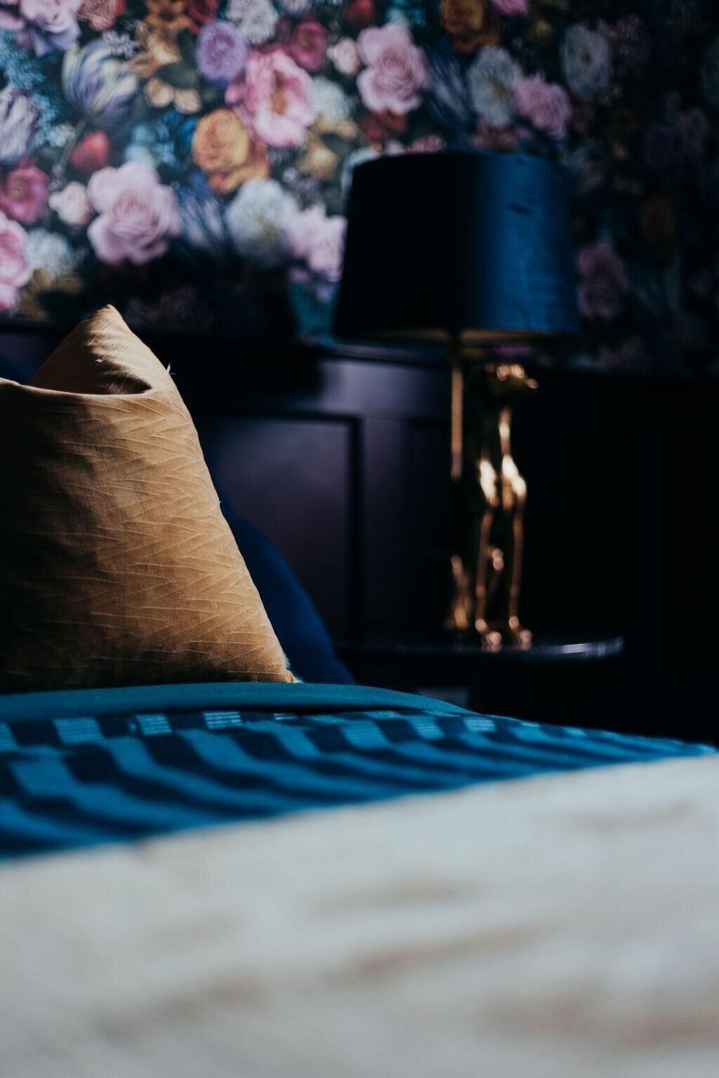 Una cama con una cubierta azul y blanca estampada, una almohada marrón y una lámpara oscura sobre una mesita de noche.