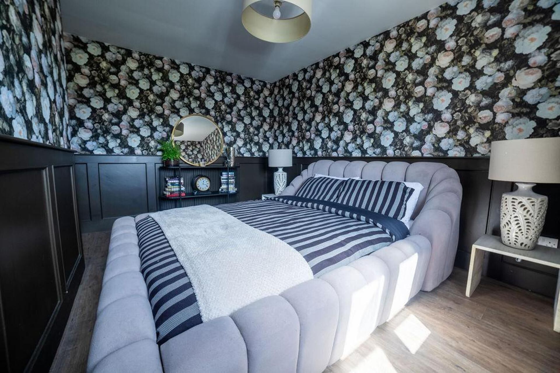 A bedroom with a large bed, a bedside table, a lamp, and a wall with a floral pattern.