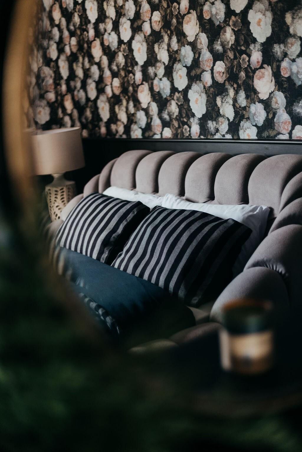 A bedroom with a tufted headboard, striped pillows, and a floral wallpaper.