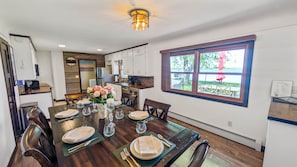 Un coin repas avec une table en bois dressée pour quatre personnes, un vase de fleurs et une vue sur l’extérieur grâce à une grande fenêtre.