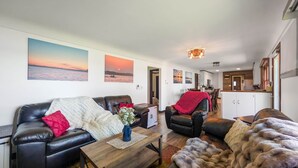 Una sala de estar con un sofá, una mesa de centro y un plaid de piel. Hay cuadros en la pared y una cocina al fondo.