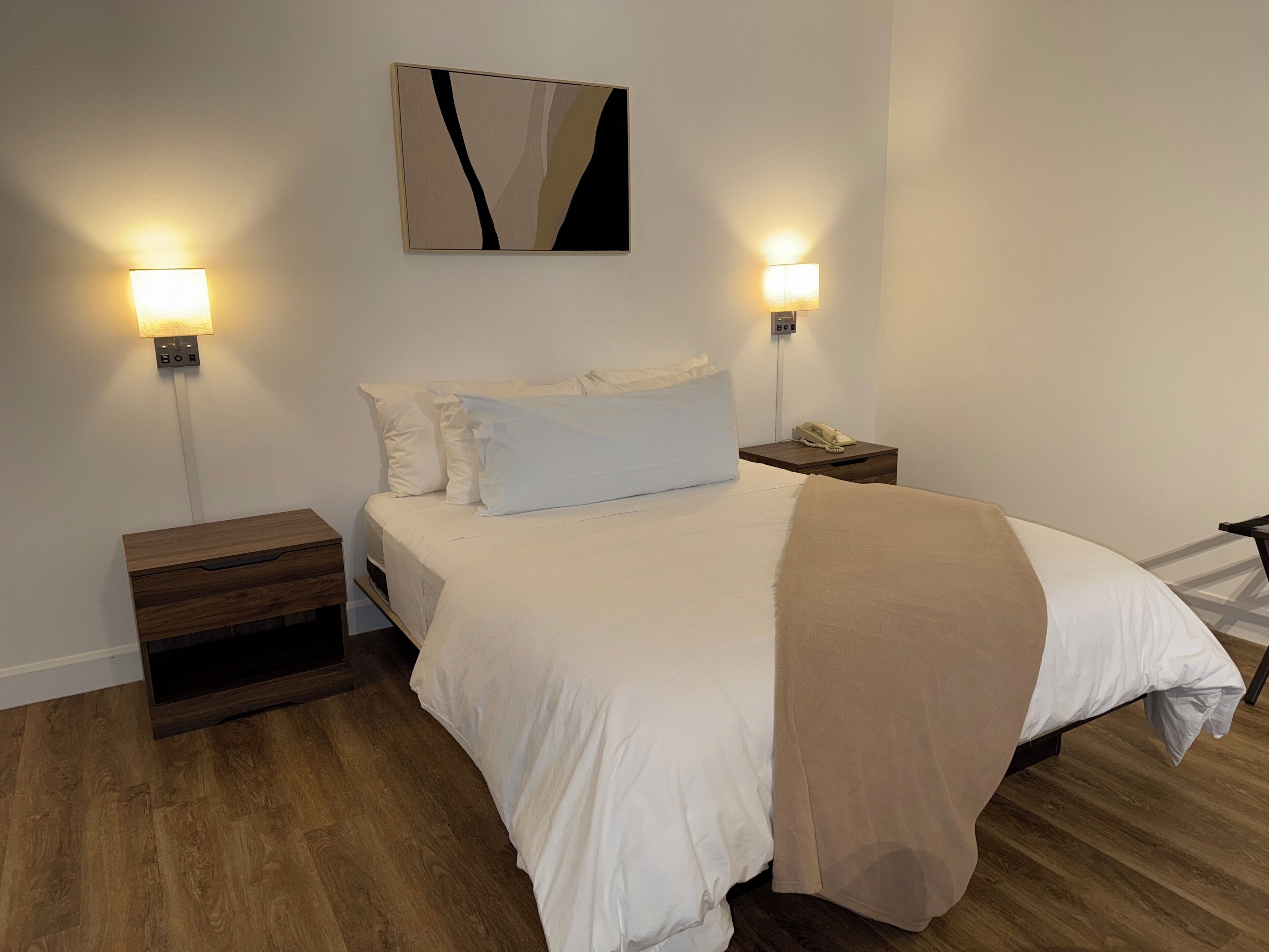 A neatly arranged hotel room with a large bed, two bedside tables, a nightstand, and a wall-mounted abstract artwork.