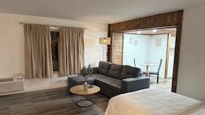 A modern bedroom with a grey sofa, a round wooden coffee table, a white bed, and a dining area in the background. - Hôtel saint-côme (Saint-Côme)