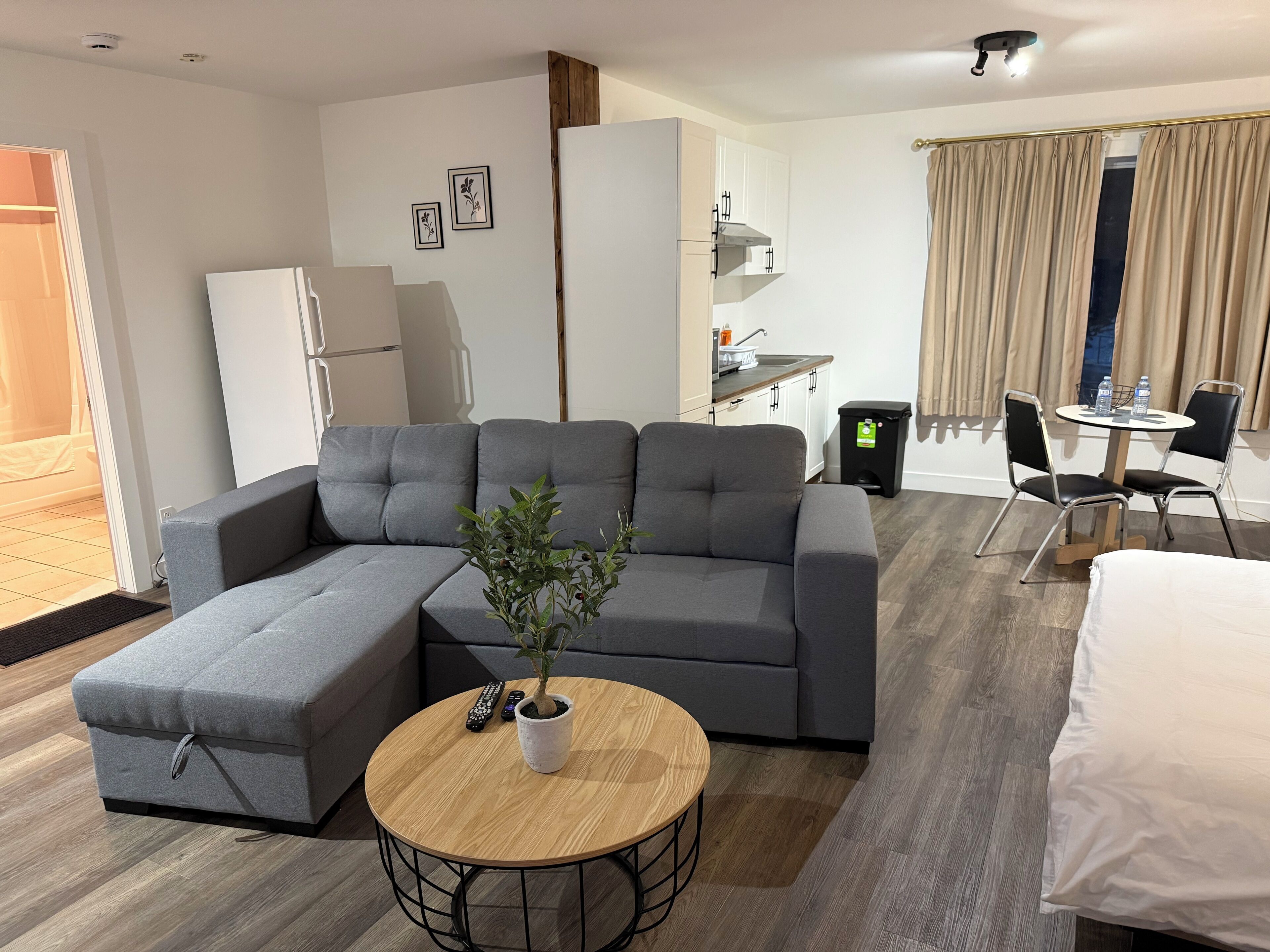 A modern living room with a grey sectional sofa, a round wooden coffee table, and a small potted plant. There is a kitchenette in the background with a refrigerator, sink, and cabinets. A dining area with a table and chairs is visible through a window with curtains.
