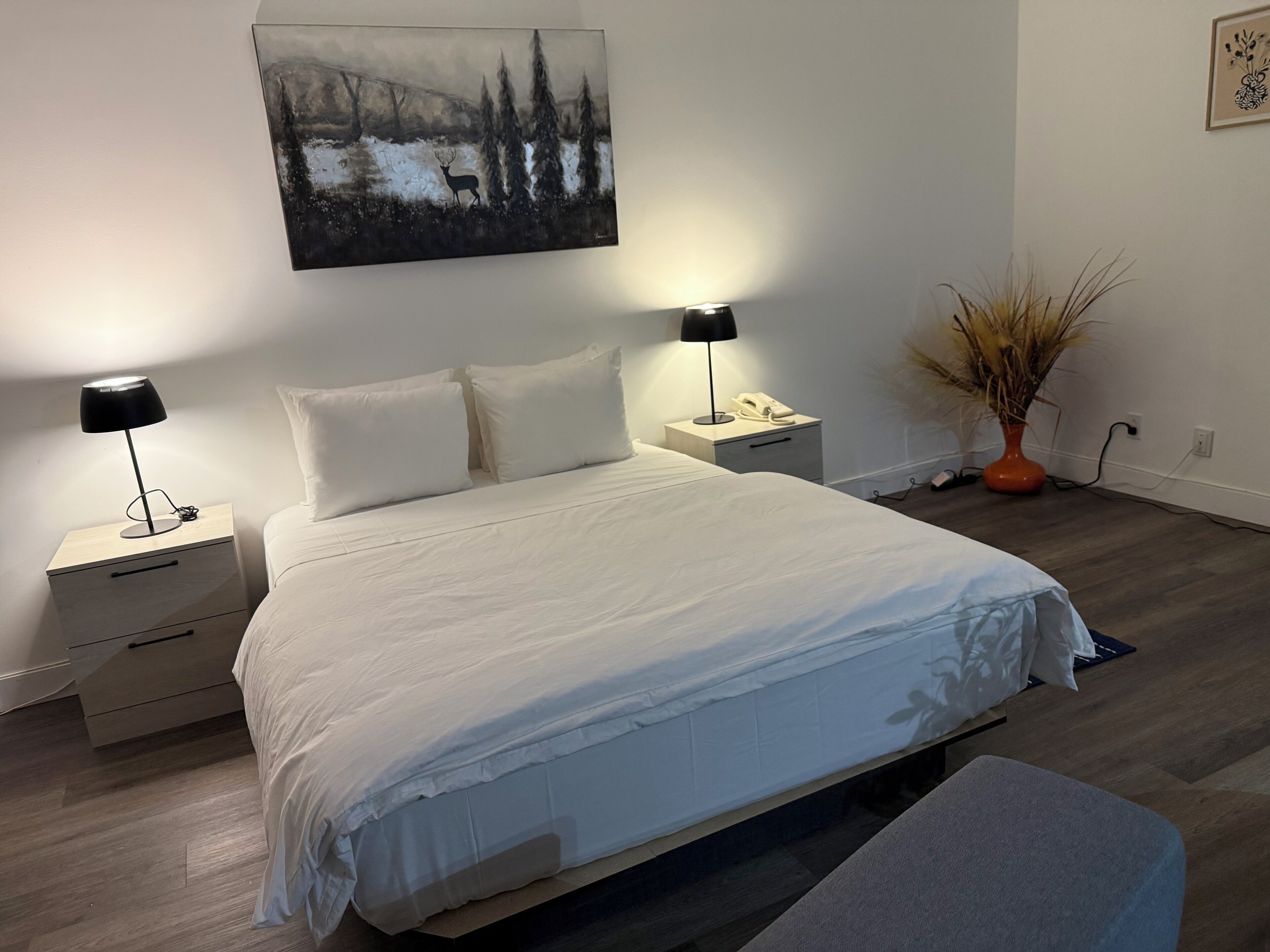 A bedroom with a large bed, two bedside tables with lamps, a nightstand, a vase with dried plants, and a painting on the wall.
