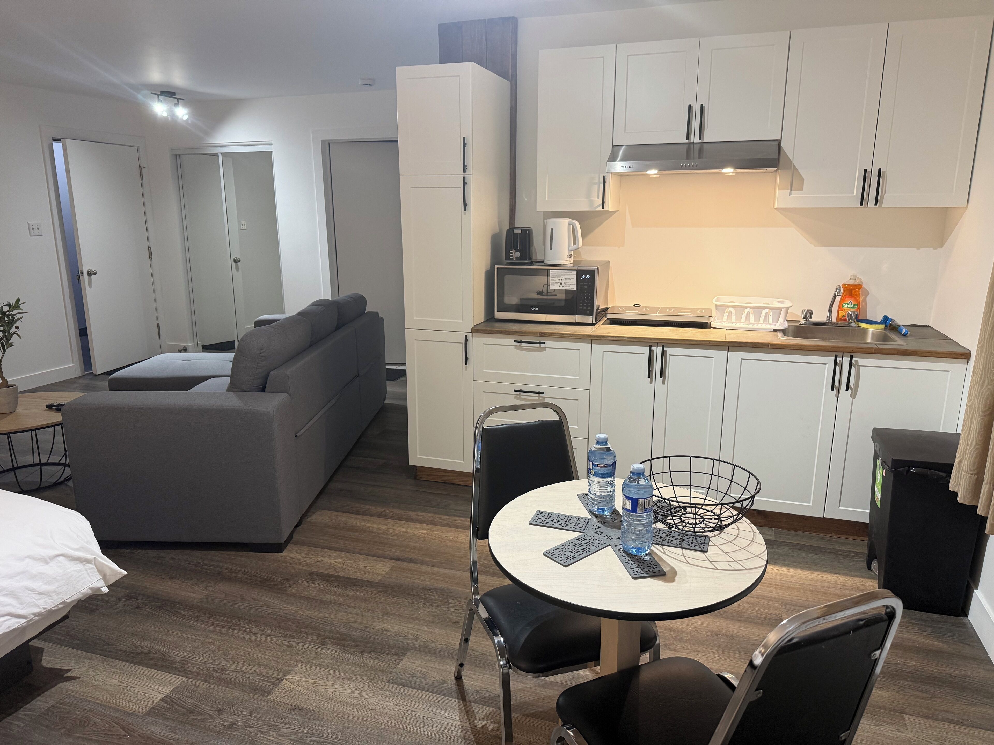 Una habitación de hotel moderna con un sofá gris, una mesa de comedor redonda con sillas, una zona de cocina con armarios blancos y un microondas.
