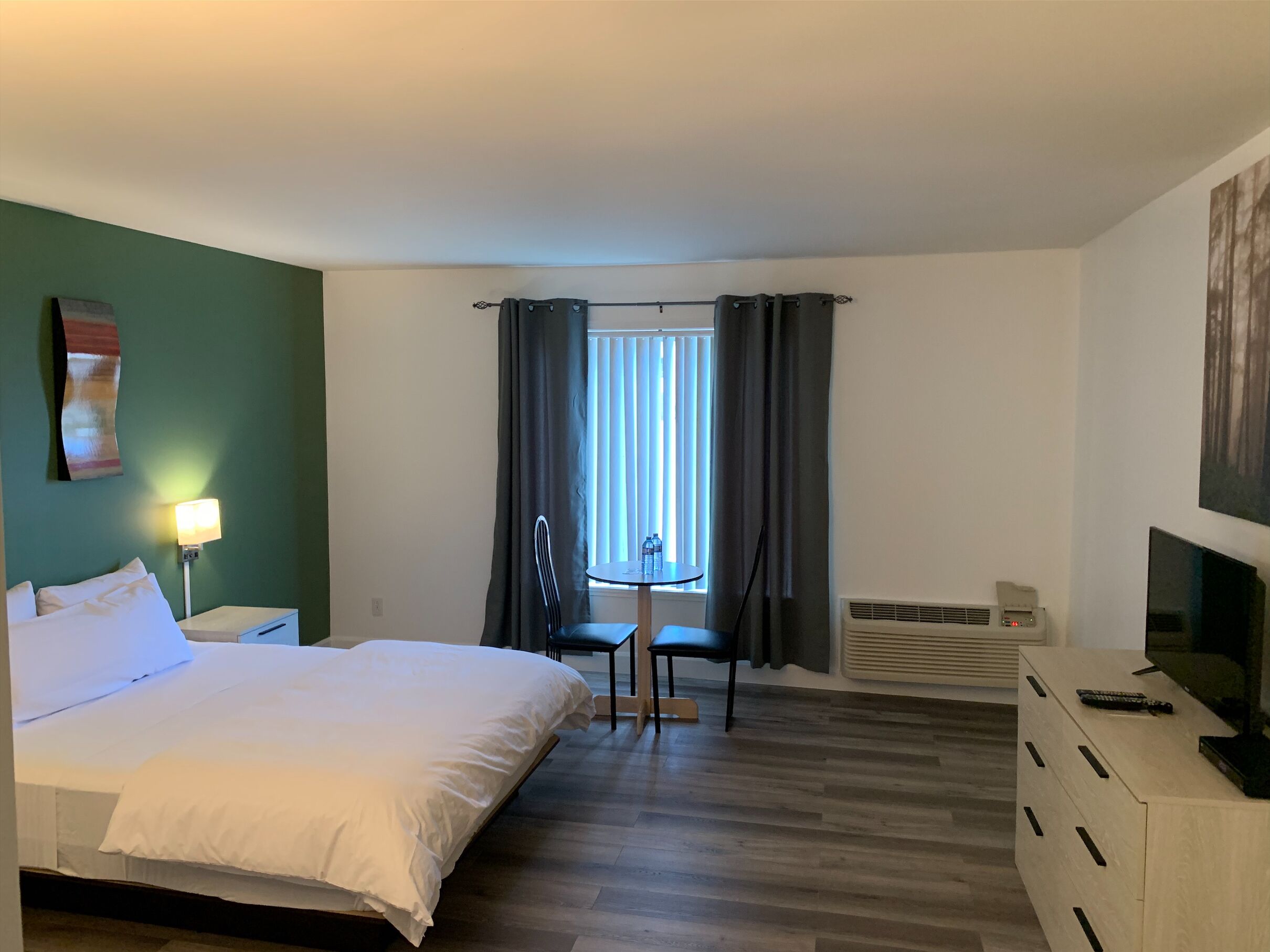 A hotel room with a bed, a desk with a TV, a chair, a table, and a window with curtains.
