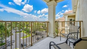 A balcony with a view of a parking lot and greenery, a table with chairs, and a building with a tiled roof. - Exclusive apartment at Vista Cay Resort (Orlando)