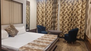 A hotel room with a large bed, two armchairs, a small round table, and curtains with a leaf pattern. - The Aura Grand (Rohtak)
