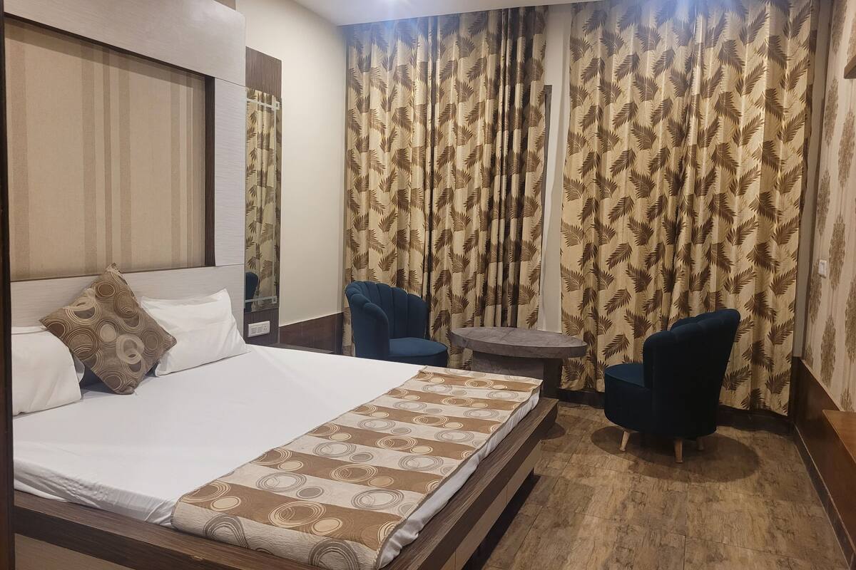 Hotellihuone, jossa on suuri sänky, kaksi nojatuolia, pieni pyöreä pöytä ja lehvikuvioilla koristellut verhot.