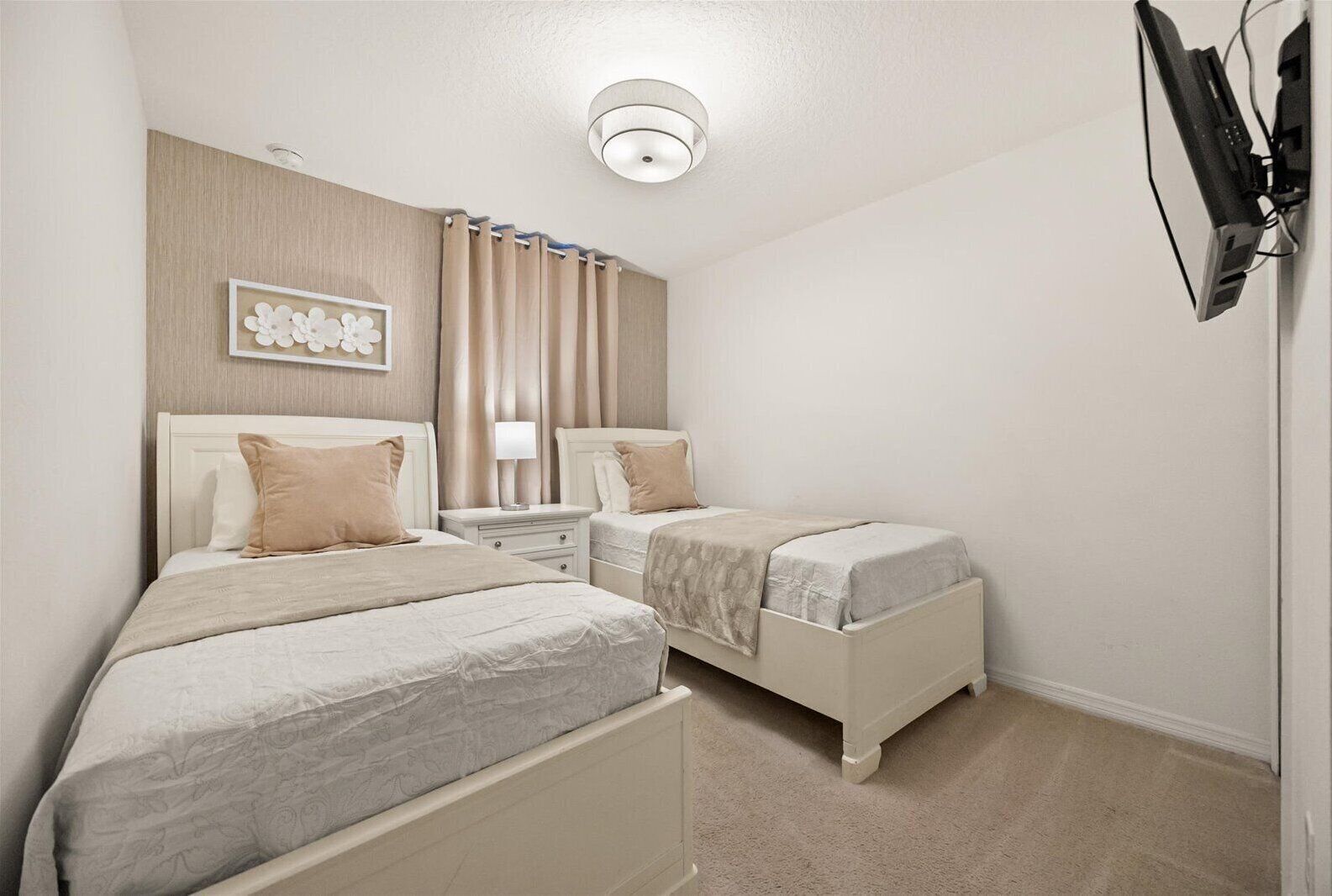 A hotel room with two beds, a bedside table with a lamp, a ceiling light, and a TV mounted on the wall.