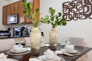 Una mesa de comedor dispuesta con platos, tazones y cubiertos blancos, y adornada con dos jarrones con follaje verde.