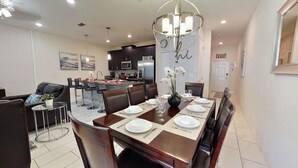 A modern dining area with a long table set for four, a kitchen in the background, and a living area with a sofa and coffee table. - Beautiful 4BR Storey Lake Resort close to Disney (Kissimmee)