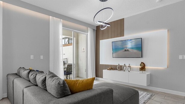 Un salon moderne avec un canapé d’angle gris, un coussin jaune, un téléviseur fixé au mur et une console blanche agrémentée d’objets décoratifs.