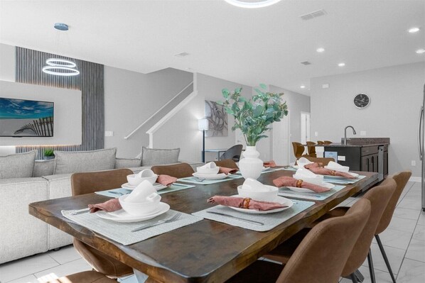 Un espace de repas moderne avec une longue table en bois dressée pour quatre personnes, un canapé gris, un escalier et, au fond, un coin cuisine.