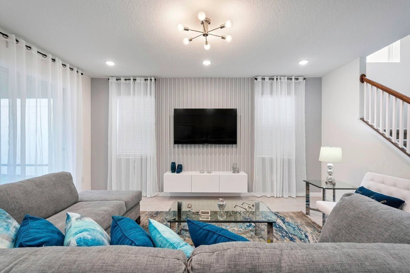 Una sala de estar moderna con un televisor de pantalla plana, cortinas blancas, una mesa de centro de vidrio y una escalera.