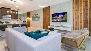 A modern living room with a sofa, a TV, and a dining area with a kitchen in the background. - Modern 6BR Home with Water Club House (Kissimmee)