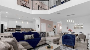 A spacious living area with a large sectional sofa, a blue armchair, and a dining table set for six. A kitchen with white cabinets and a microwave is visible in the background.