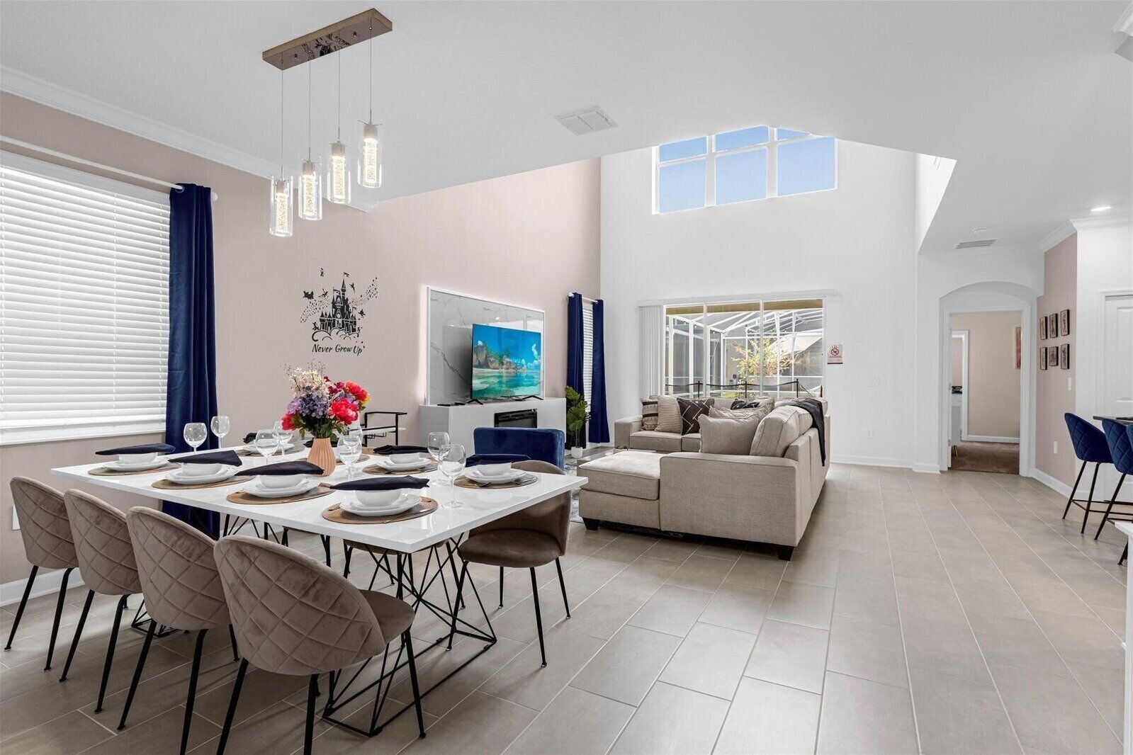 Un espace repas moderne avec une table blanche dressée pour quatre personnes, un canapé beige et un téléviseur fixé au mur.