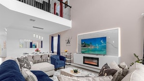 A modern living room with a fireplace, a large TV, and a dining area in the background.