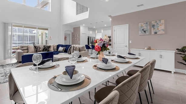 A spacious dining area with a long table set for a meal, a kitchen in the background, and a high ceiling with natural light.