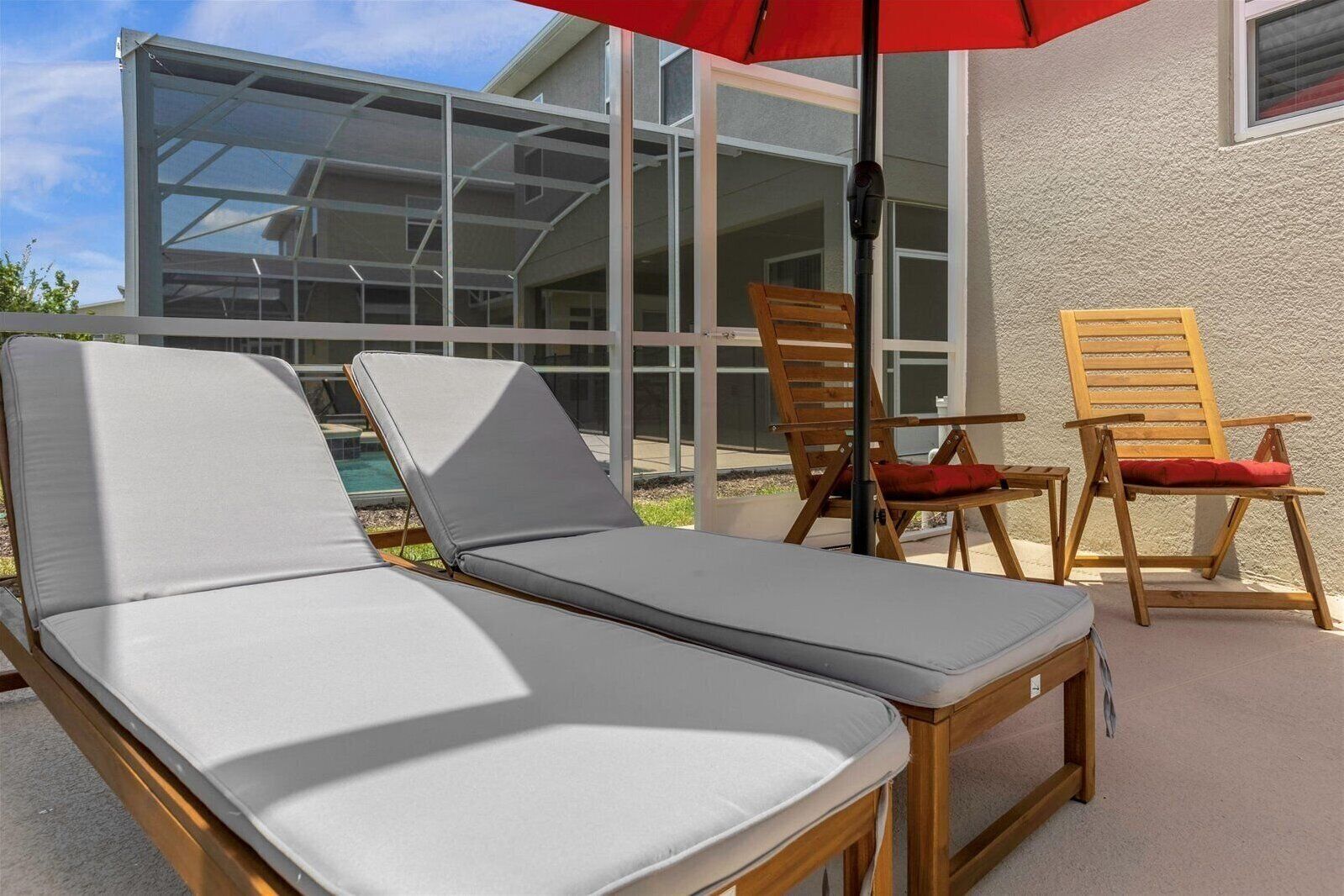 Eine Sonnenliege und zwei Holzsessel mit Kissen, platziert auf einer Terrasse mit einem Pool im Hintergrund.