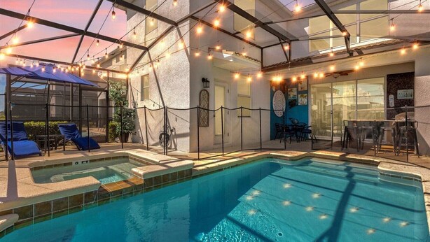 A swimming pool with lounge chairs and a covered seating area, surrounded by a fence and string lights.
