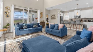 A spacious living area with two sofas and a coffee table, a kitchen with modern appliances, and a large window offering a view of the outdoors.