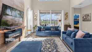 A spacious living room with a large TV, a fireplace, and a view of the outdoors through large windows.