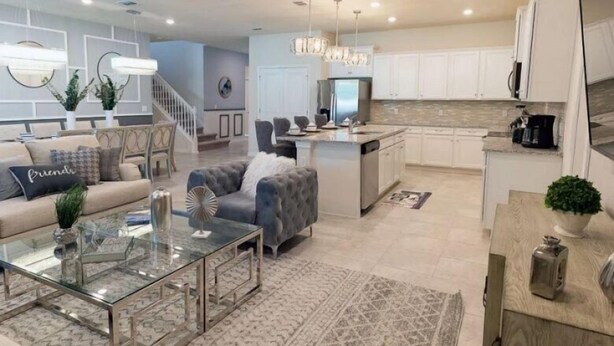 Una sala de estar moderna con una mesa de centro de vidrio, un sofá con almohadones decorativos y un sillón gris. La cocina cuenta con gabinetes blancos, un refrigerador de acero inoxidable y una isla central con un lavamanos.