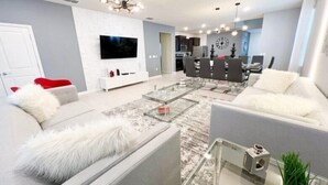 A modern living room with a sofa, coffee table, and TV. There is a dining area with chairs and a table. The room has a neutral color scheme and modern decor.