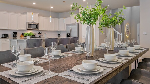 Un comedor moderno con una larga mesa de madera dispuesta para una comida, rodeado de armarios blancos y una cocina al fondo.