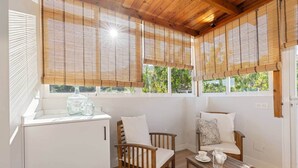 Ein gemütliches Zimmer mit Holzbalkendecke, einem kleinen Tisch mit Krug und Tassen, zwei Holzstühlen mit Kissen, einem weißen Schrank und Bambusrollos an den Fenstern.