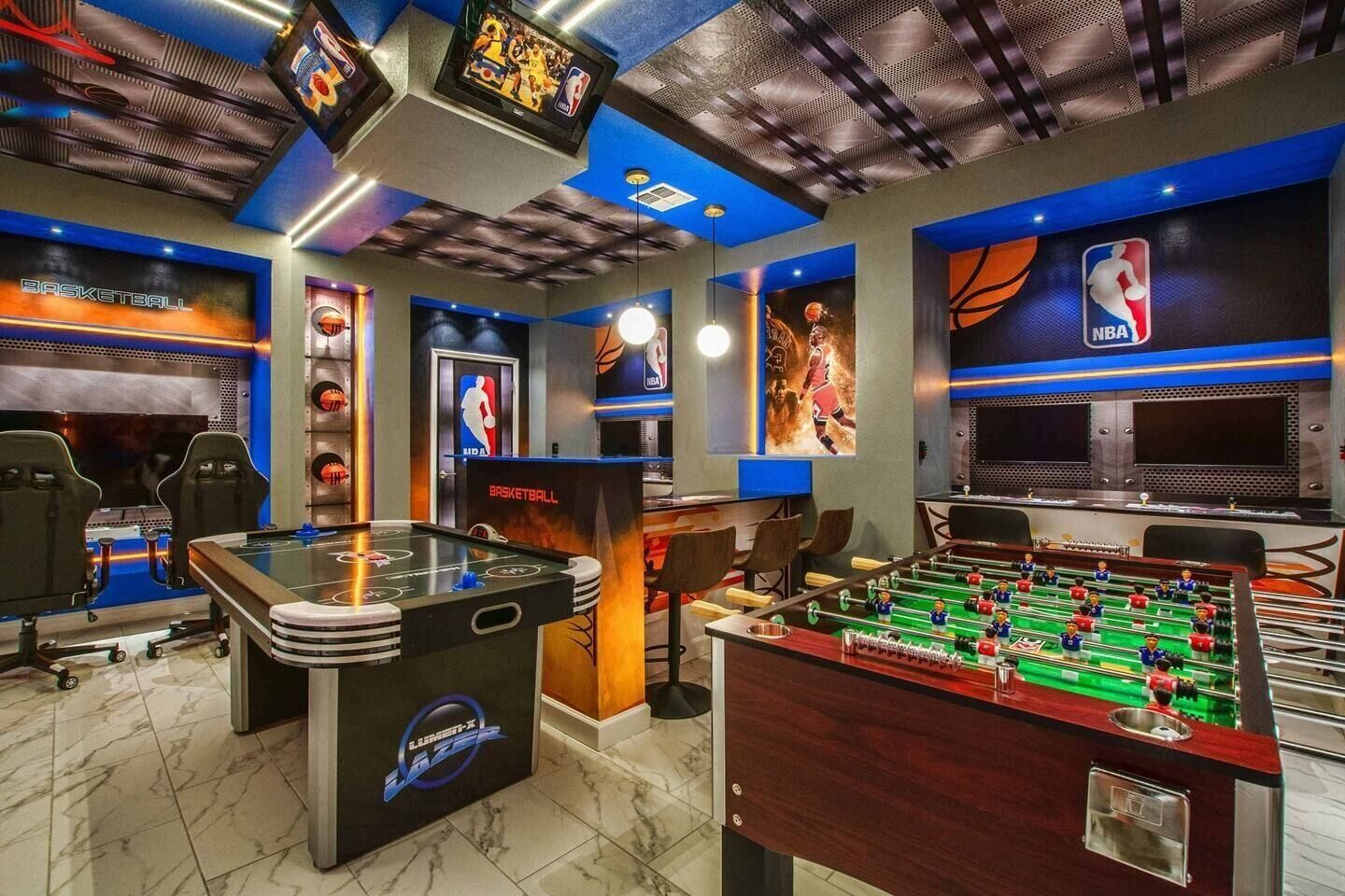 A room with a foosball table, a basketball table, and a large screen displaying a basketball game.
