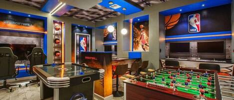 A room with a foosball table, a basketball table, and a large screen displaying a basketball game.