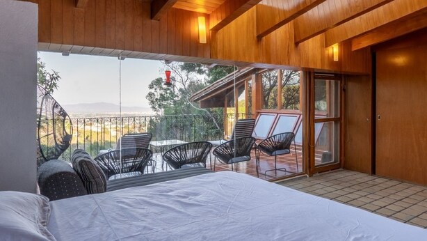 A bedroom with a large bed, a balcony with chairs, and a view of the outdoors.