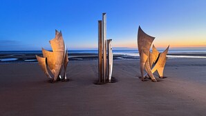 Drie abstracte metalen sculpturen op een strand, met de oceaan op de achtergrond.
